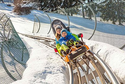 Zwei Kindern rodeln mit dem Alpsee Coaster der Alpsee Bergwelt bei Immenstadt im Winter den Berg hinunter.