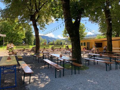 Ein einladender Außenbereich mit mehreren Holztischen und Bänken unter Bäumen. Im Hintergrund sind die Berge und ein gelbes Gebäude zu sehen.
