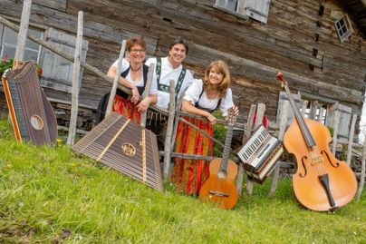 Drei Musiker stehen vor einer alten Holzscheune und lächeln. Verschiedene traditionelle Musikinstrumente liegen vor ihnen auf dem Boden.