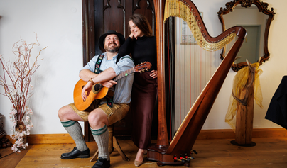 Ein Mann spielt Gitarre und lächelt, während eine Frau neben ihm steht. Im Hintergrund ist eine Harfe zu sehen, die auf dem Boden steht.