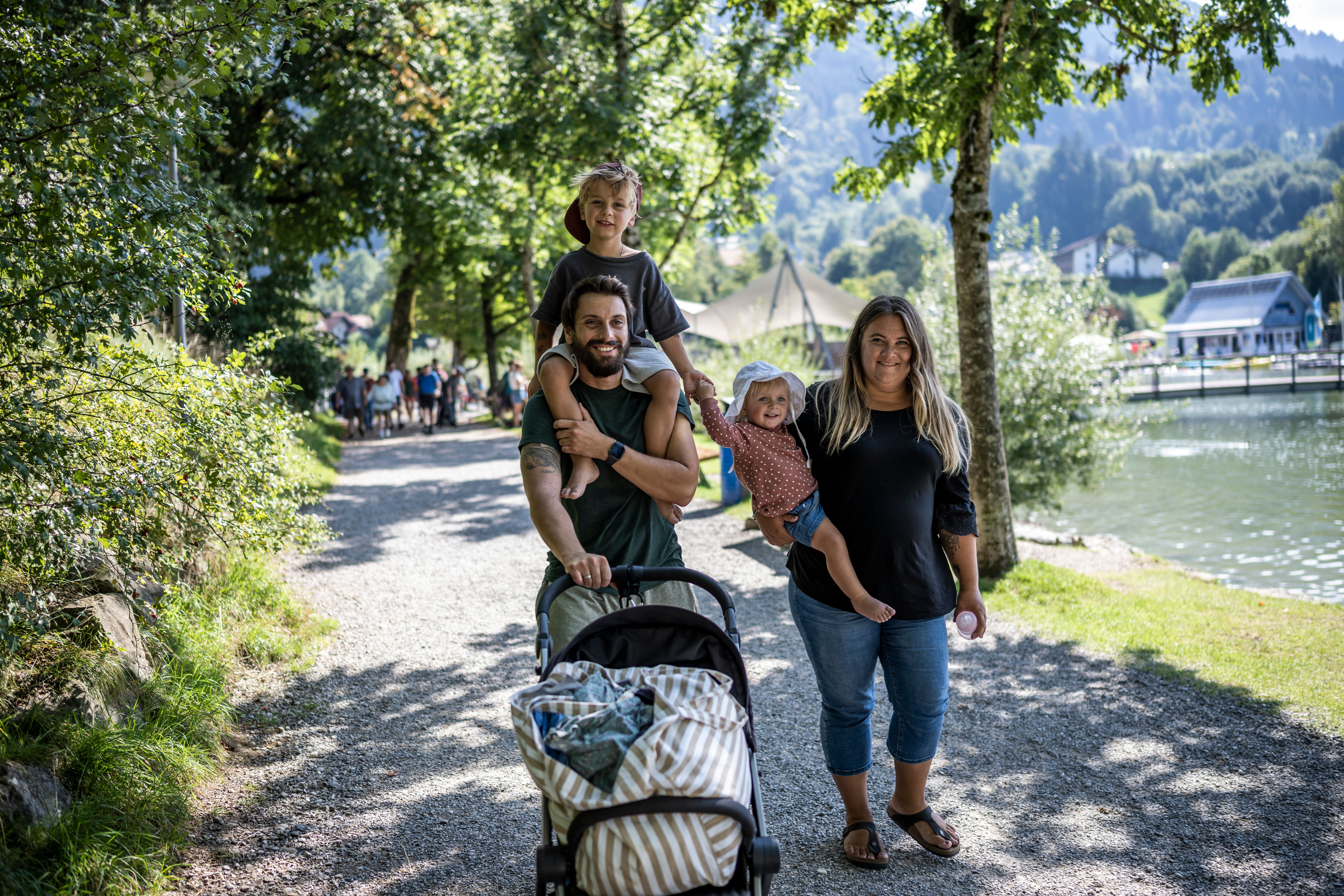 Eine Familie läuft mit Ihrem Kinderwagen entlang der Seepromenade am Alpsee in Immenstadt. Der Papa trägt den Jungen auf den Schultern und die Mama das kleine Mädchen auf ihren Armen.