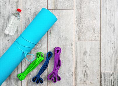 Eine blaue Yogamatte liegt auf einem hellen Boden, daneben liegen Gummibänder in verschiedenen Farben und eine Flasche Wasser. Ideal für das Fitness-Workout zu Hause.
