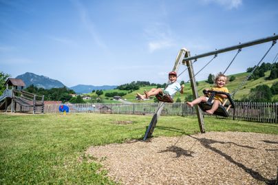 Zwei Kinder schwingen fröhlich auf einer Schaukel im Freien. Im Hintergrund sind grüne Wiesen und Berge sichtbar.