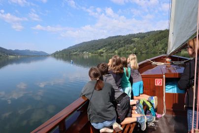 Eine Gruppe von Kindern steht auf einem Boot und schaut auf einen ruhigen See. Die Landschaft ist grün und die Himmel ist blau mit einigen Wolken.