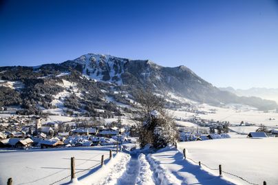 Das Bild zeigt von leicht erhöhter Position den Ort Rettenberg im Winter. Im Hintergrund ist die Breitseite des Bergs Grünten zu sehen.