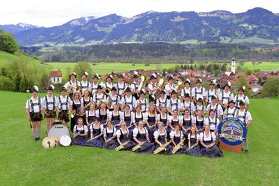 Eine große Gruppe von Musikern in traditionellen Trachten steht auf einer Wiese. Im Hintergrund sind Berge und ein Dorf zu sehen.