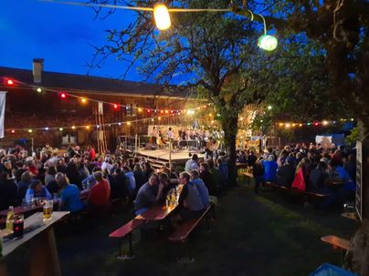 Eine lebhafte Veranstaltung im Freien mit vielen Menschen, die an langen Tischen sitzen. Auf einer Bühne spielt eine Band, während bunte Lichter die Atmosphäre erhellen.