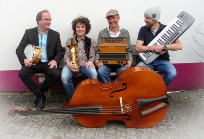 Eine Band sitzt zusammen und spielt verschiedene Instrumente. Im Vordergrund stehen ein Cello und mehrere Musiker mit einer Trompete, einem Keyboard und anderen Musikinstrumenten.
