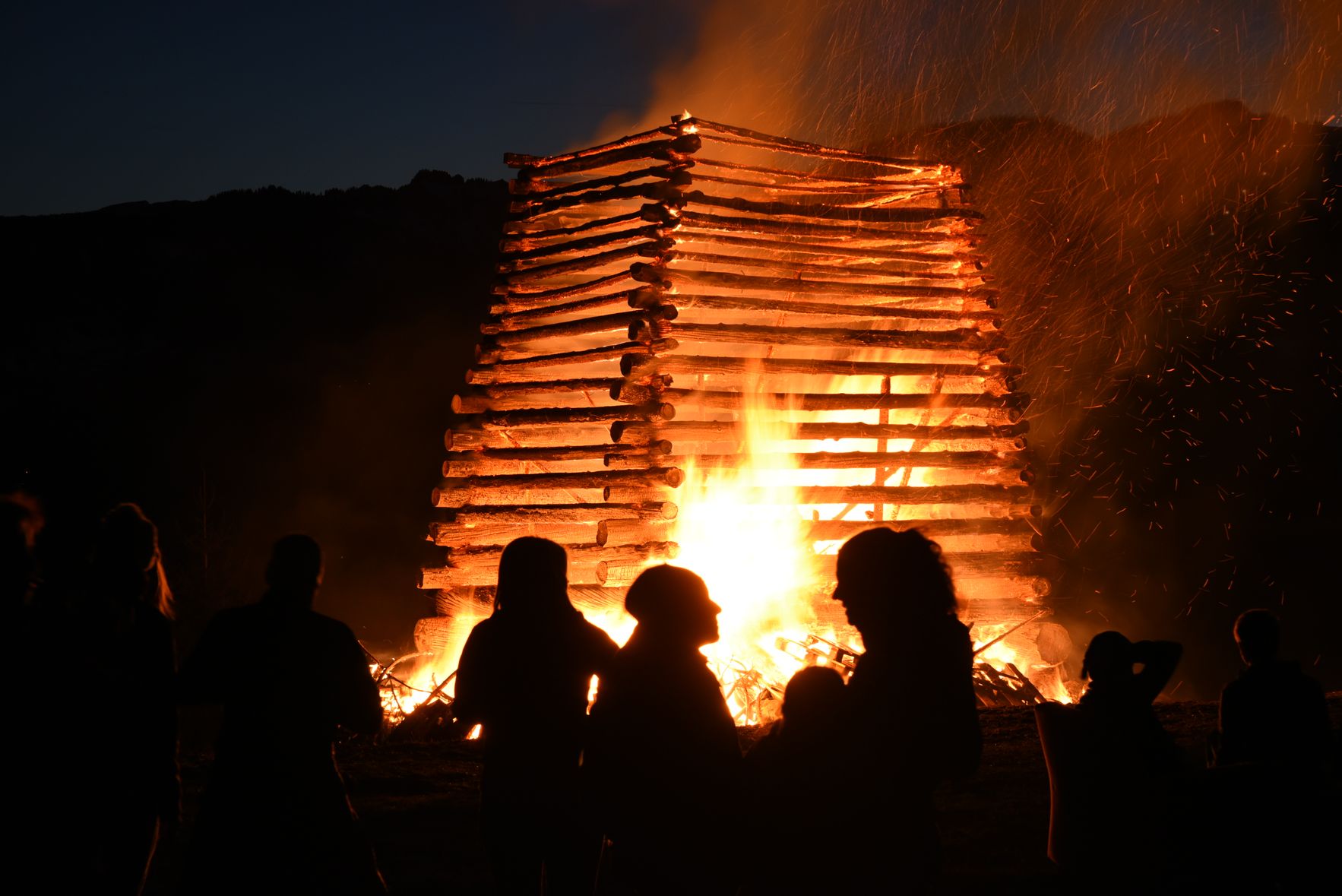 Auf dem Bild sieht man den brennenden Funken in Altstädten. Die Flammen lodern hell. Davor sieht man drei Personen, die sich unterhalten und den Funken ansehen.