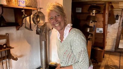 Eine lächelnde Frau steht in einer gemütlichen Küche und bereitet etwas vor. Sie trägt ein helles Oberteil und hat blonde Haare, die sie zusammengebunden hat.