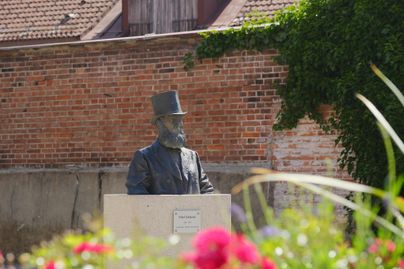 Eine Statue eines Mannes mit einem Zylinderhut und einem Bart steht in einer Gartenumgebung. Im Hintergrund sind Backsteinmauern sichtbar.