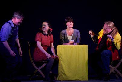 Eine Theateraufführung mit vier Schauspielern, die um einen Tisch sitzen. Die Atmosphäre ist lebhaft und jeder Schauspieler zeigt eine eigene Emotion.