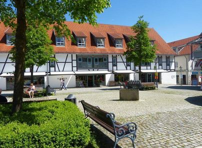 Ein malerischer Platz mit Fachwerkhäusern und grünen Bäumen. Menschen sitzen auf Bänken und genießen die sonnige Atmosphäre.