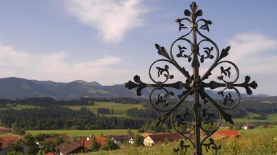 Ein kunstvoller Eisenkreuz steht auf einer Wiese mit Blick auf grüne Hügel und ein kleines Dorf. Der Himmel ist blau mit einigen Wolken.