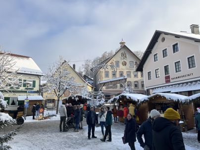 Ein winterlicher Marktplatz mit schneebedeckten Gebäuden und festlich dekorierten Ständen. Menschen schlendern durch die verschneite Umgebung und genießen die Atmosphäre.