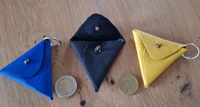 Drei kleine, dreieckige Schlüsselanhänger in Blau, Schwarz und Gelb liegen auf einem Holztisch. Neben den Anhängern befinden sich zwei Münzen.