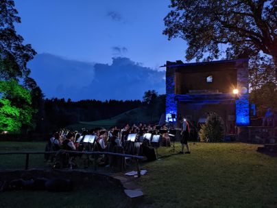 Ein Orchester spielt bei Dämmerung in einer schönen Umgebung. Im Hintergrund ist ein beleuchtetes Gebäude und ein klarer Himmel zu sehen.
