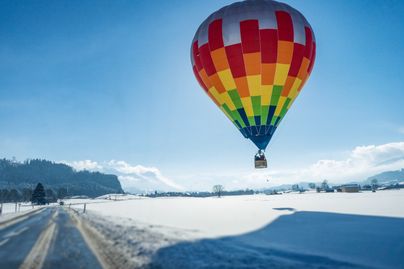 Auf einer schneebedeckten Wiese, neben der Zufahrtsstraße in Richtung Agathazell, setzt ein bunter Heißluftballon im Winter zur Landung an. Agathazell ist ein ein Ortsteil der Gemeinde Burgberg am Fuße des Grüntens.