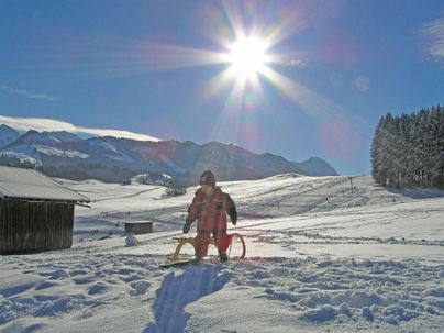 Ein Junge steht in einer schneebedeckten Landschaft mit Bergen im Hintergrund. Die Sonne scheint hell am klaren Himmel.