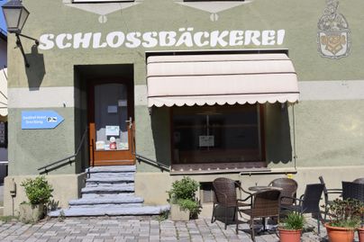Eine Bäckerei mit dem Namen "Schlossbäckerei" und einem klaren Eingang. Vor der Bäckerei stehen Tische und Stühle auf dem Pflaster.