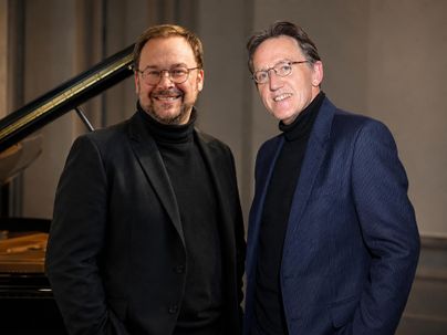 Zwei Männer stehen lächelnd nebeneinander vor einem Klavier. Beide tragen schwarze oder dunkelblaue Kleidung und haben einen entspannten Ausdruck.