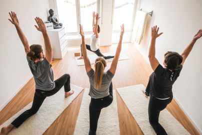 Eine Gruppe von Frauen praktiziert Yoga in einem hellen Raum. Sie strecken die Arme nach oben und konzentrieren sich auf ihre Übungen.