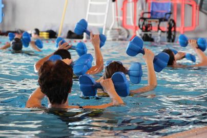 Eine Gruppe von Menschen trainiert im Wasser mit blauen Hanteln. Es ist eine Aquafitness-Klasse in einem Hallenbad.