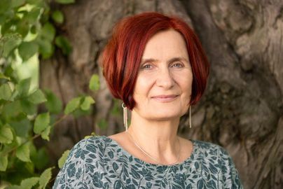 Eine Frau mit rotem Haar trägt ein blumiges Oberteil und lächelt in die Kamera. Im Hintergrund sind grüne Blätter und ein Baumstamm sichtbar.