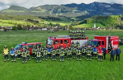 Eine Gruppe von Feuerwehrleuten steht in einem grünen Feld mit Bergen im Hintergrund. Im Vordergrund sind mehrere Feuerwehrautos zu sehen.