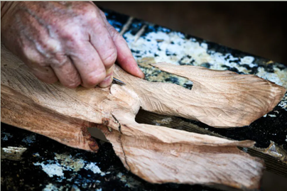 Eine handwerkliche Holzschnitzerei mit einer Hand, die ein Stück Holz bearbeitet. Die Holzoberfläche zeigt feine Details und Strukturen.