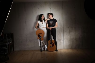 Ein Paar steht in einem Studio vor einer Betonsäule. Beide halten Akustikgitarren und lächeln sich an.