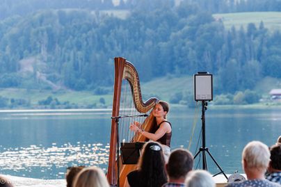 Eine Harfenistin spielt am Ufer eines Sees. Im Hintergrund sind Bäume und Berglandschaften zu sehen.