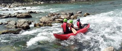 Drei Personen paddeln in einem roten Kajak auf einem rauschenden Fluss. Die Umgebung ist felsig und grün, und es fließt viel Wasser.