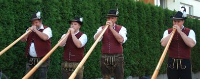 Vier Männer in traditioneller Tracht spielen auf langen Alphörnern. Im Hintergrund ist eine grüne Hecke zu sehen.