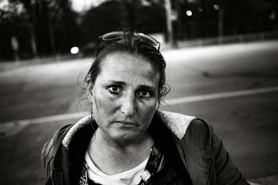 Ein Porträt einer Frau mit ernstem Ausdruck. Sie sitzt an einem Straßenrand in einer urbanen Umgebung.