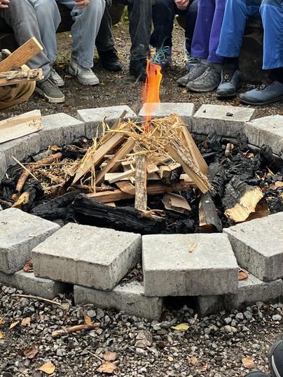 Ein Lagerfeuer brennt in einem runden Steinkreis. Um das Feuer sitzen Menschen in gemütlicher Runde.