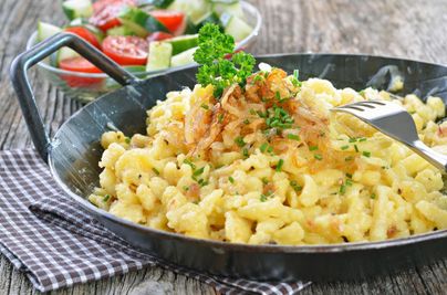 Eine leckere Portion Käsespätzle mit Röstzwiebeln und frischen Kräutern. Im Hintergrund ein frischer Salat aus Tomaten und Gurken.