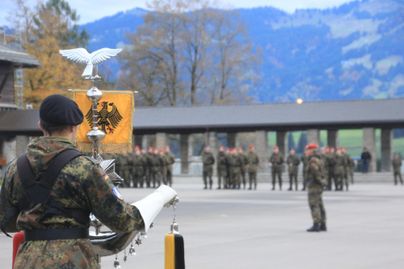 Eine militärische Zeremonie mit Soldaten in Uniform und einer Fahne im Vordergrund. Die Umgebung ist ländlich mit Bergen im Hintergrund.