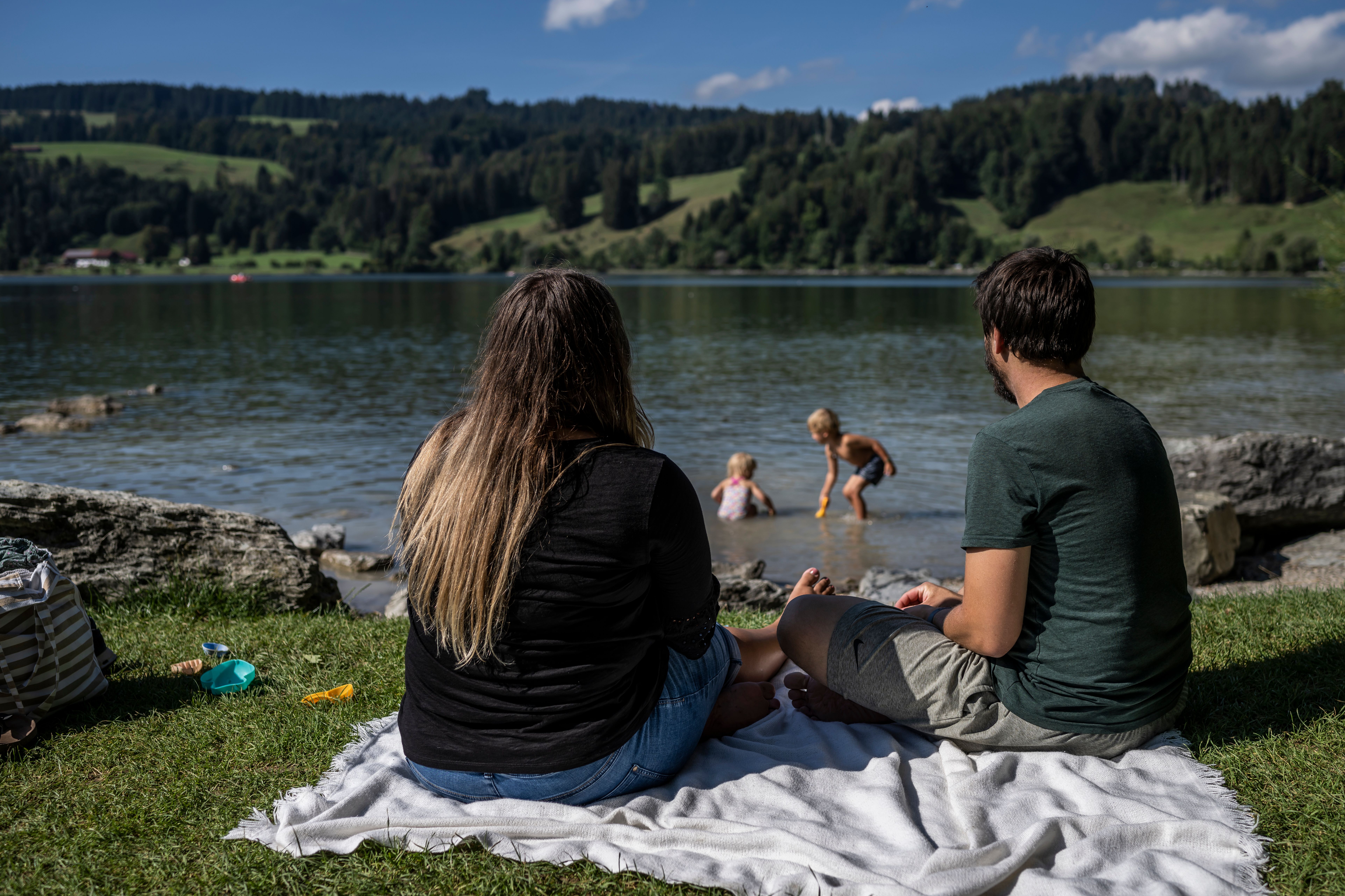 Die Eltern sitzen auf einem Handtuch am Ufer des Alpsees in Immenstadt, während die zwei Kinder im See plantschen.