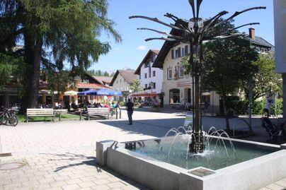 Eine hübsche Platzansicht mit einem Brunnen in der Mitte. Rundherum befinden sich Cafés und Bäume unter einem klaren blauen Himmel.