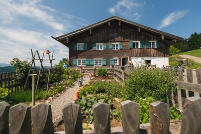 Ein traditionelles Holzhaus mit grünen Fensterläden und einem schönen Garten. Im Vordergrund befindet sich ein niedriger Zaun aus Holz.