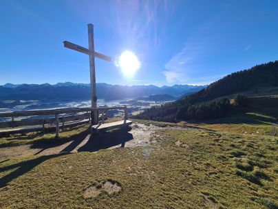 Ein schöner Ausblick von einem Hügel mit einem Kreuz. Die Sonne scheint über den Bergen und das Tal ist in sanftem Licht gehüllt.
