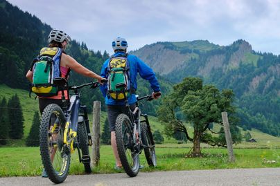 Zwei Mountainbiker stehen mit ihren E-Bikes am Stacheldrahtzaun einer Weide. Sie blicken in Richtung eines Berges im Gunzesrieder Tal.