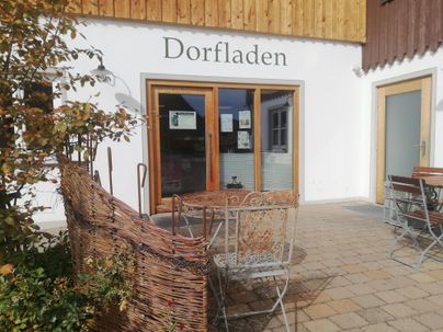 Ein Dorfaden mit großen Fenstern und einer einladenden Terrasse. Es gibt Sitzgelegenheiten aus Metall und einen schönen Blick auf die Umgebung.