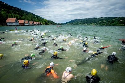 Eine Gruppe von Schwimmern nimmt an einem Wettkampf im Wasser teil. Im Hintergrund sind Häuser und eine grüne Landschaft zu sehen.