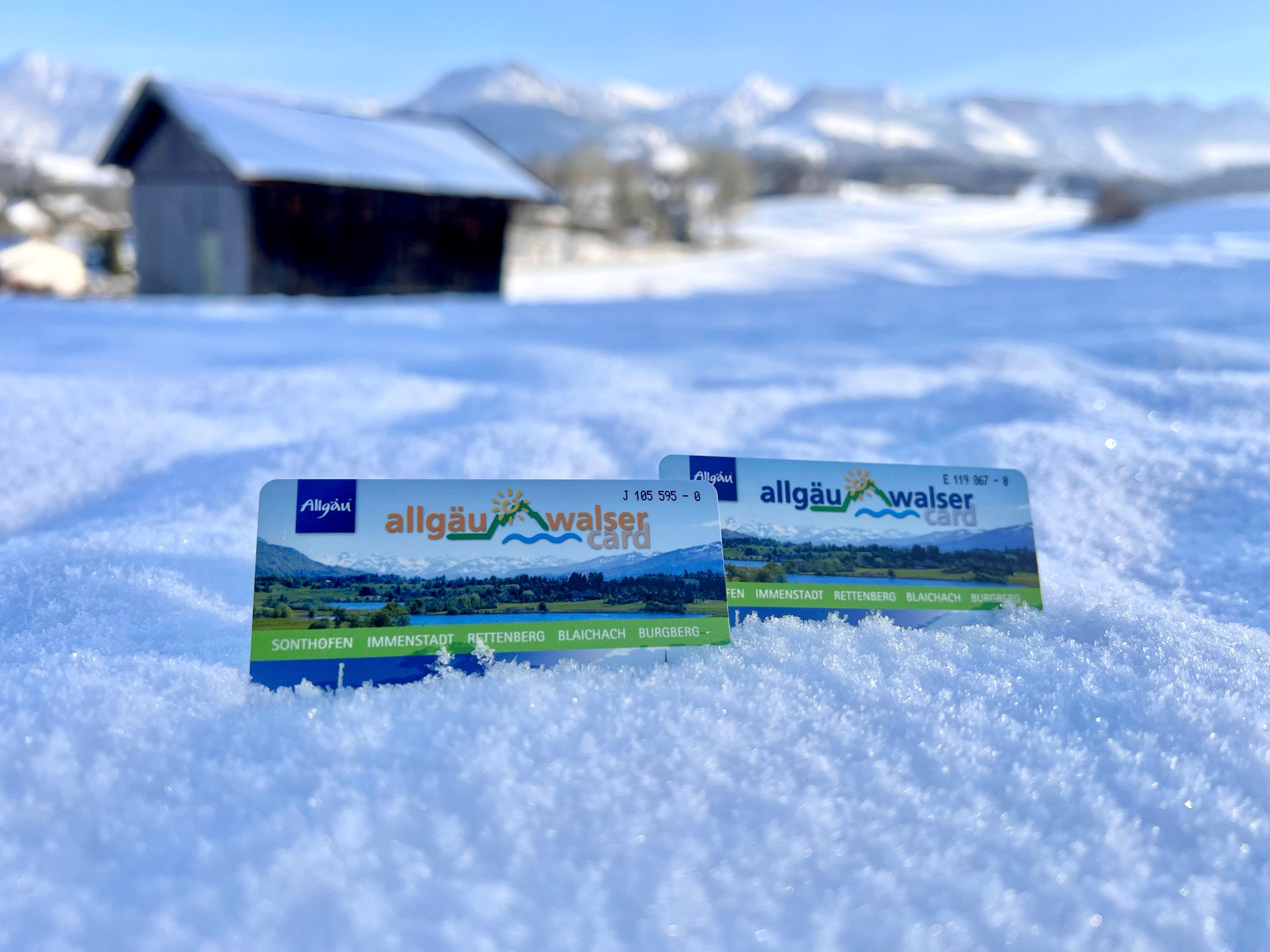Im Vordergrund sind zwei Allgäu-Walser-Cards der Alpsee-Grünten Region im Schnee zu sehen. Im Hintergrund befindet sich eine schneebedeckte Hütte und die Allgäuer Alpen.