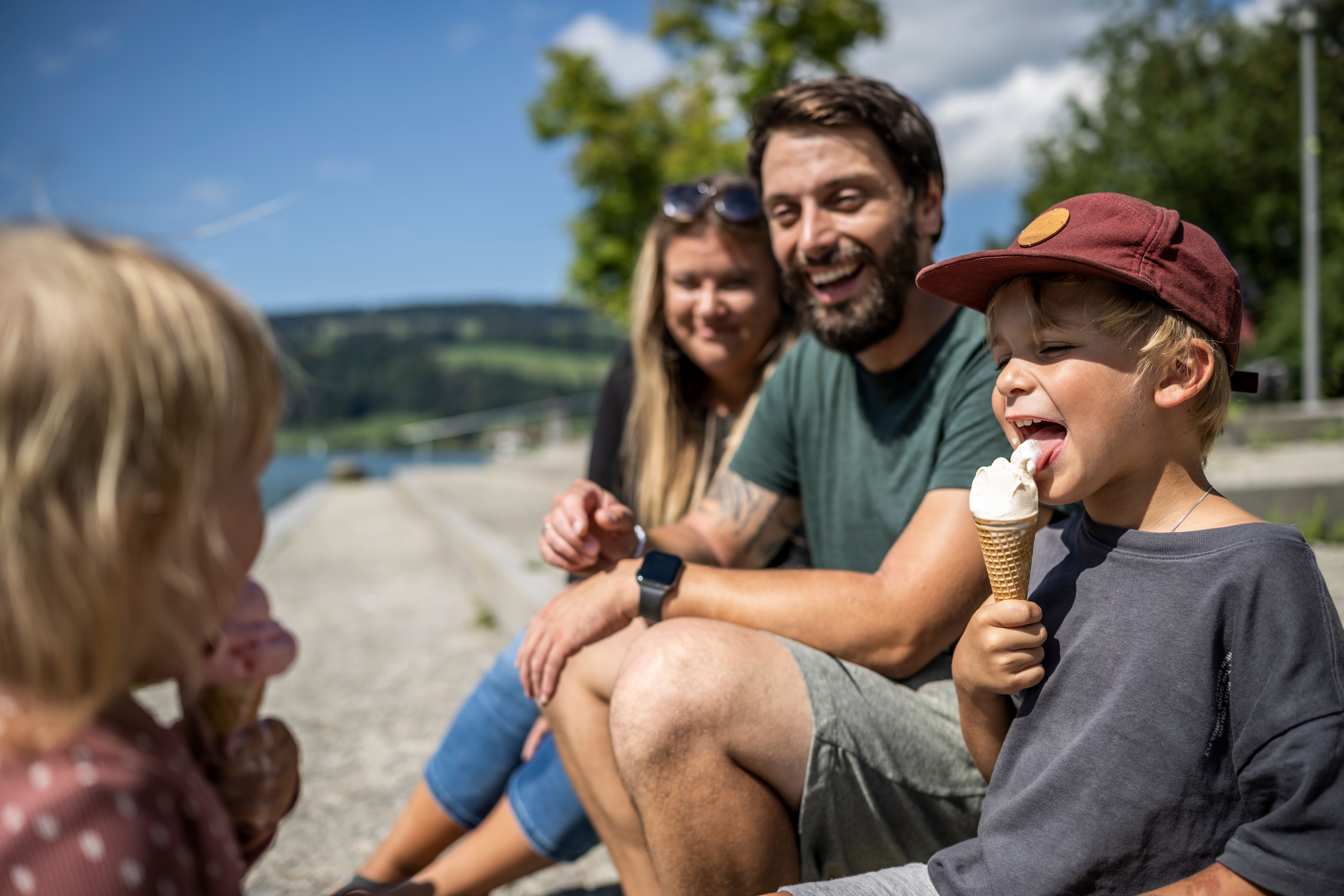 Eine Familie mit Mama, Papa und zwei Kindern sitzen an der Alpsee Promenade und lachen gemeinsam. Die zwei Kinder essen eine Kugel Eis auf der Waffel.