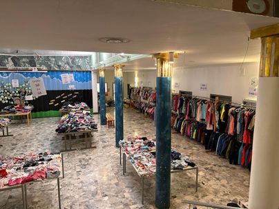 Ein großer Raum mit Tischen, die mit Kleidung bedeckt sind. An den Wänden hängen weitere Kleidungsstücke auf Kleiderständern.