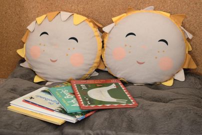Zwei lustige Kissen in Sonnenform liegen auf einem grauen Stoff. Daneben sind einige bunte Kinderbücher gestapelt.
