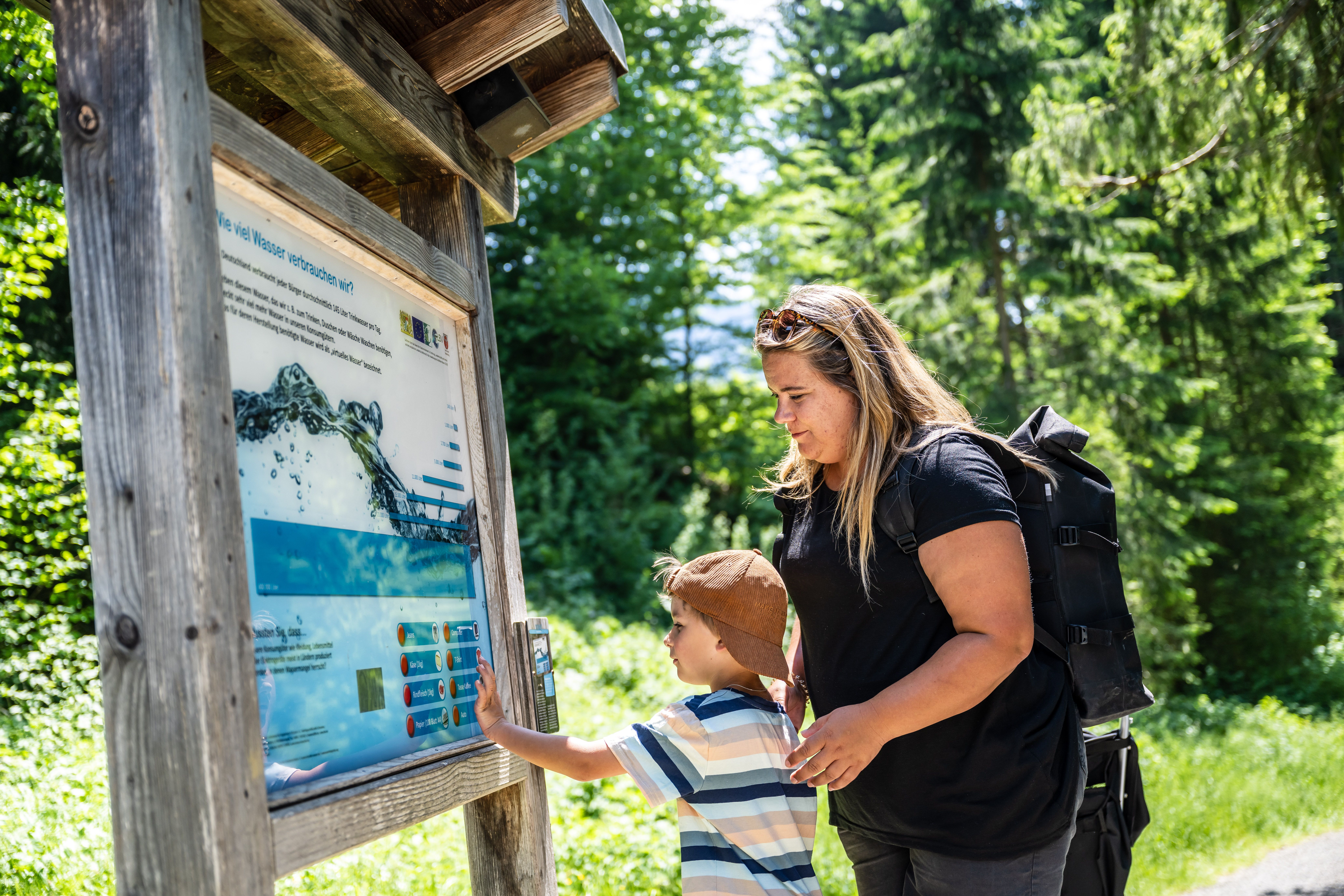 Ein Junge und seine Mutter schauen sich eine Info-Tafel zum Thema Wasser entlang des Erlebnispfad am Glatschbach an.